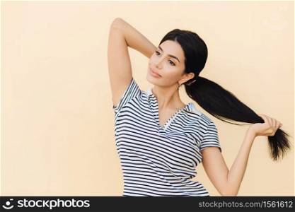 Pensive dark haired female makes pony tail, has full lips and pleasant appearance, wears casual outfit, stands against studio background, focused into distance. People, beauty and lifestyle concept