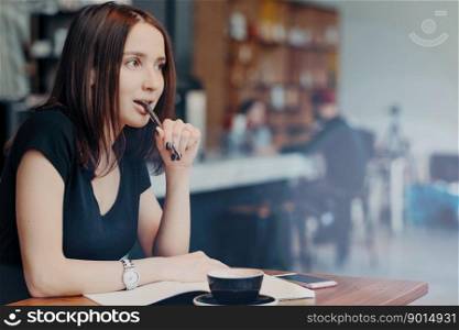 Pensive female blogger writes in notepad ideas for publication in blog, sits against cafe interior, keeps pen at mouth, drinks coffee focused away, dressed in black t shirt, thinks about plans