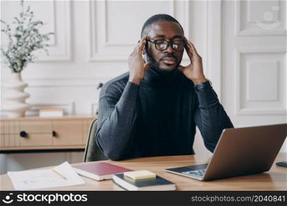 Pensive focused young African male bookkeeper sitting at desk at home office in front of laptop eyes closed fingers gently touching temples during hard cogitative process while working remotely online. African male bookkeeper sits at desk in front of laptop eyes closed fingers gently touching temples