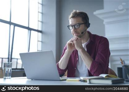 Pensive German businessman in headphones sits at desk while remotely working from home looks at laptop display with concentration, hands folded under his chin when reads sales report from main office. Pensive German businessman in headphones sits at desk while remotely working from home