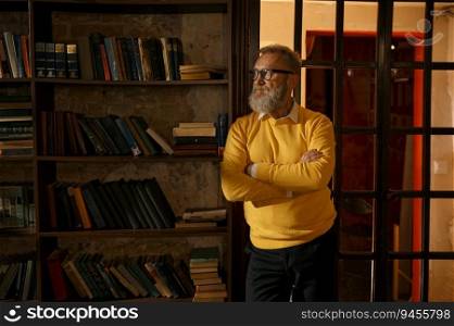 Pensive gray-haired senior man standing with arms crossed on chest at home library aristocratic interior. Mature older man dreaming about something leaned at bookshelves. Pensive senior man standing with arms crossed on chest at home library