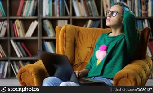 Pensive hipster girl in stylish clothes and trendy specatcles concentrated on generating creative ideas while sitting in cozy chair in library. Pondering talented female freelancer working on project using laptop pc over bookshelves background.