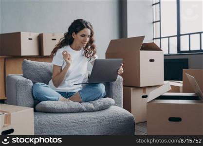 Pensive hispanic woman sits with cardboard boxes uses laptop makes order for transport services on moving day, buys furniture for new home, searching repairs company or interior design ideas.. Hispanic girl sits with cardboard boxes uses laptop makes order for transport services on moving day