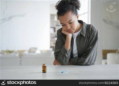 Pensive ill young mixed race girl with chronic disease looks at medical pills, medicines before taking meds, sitting at table at home. Unhealthy teen lady doubts, pondering whether to take medication.. Pensive mixed race girl looks at medical pills, medicines before taking meds, sitting at table