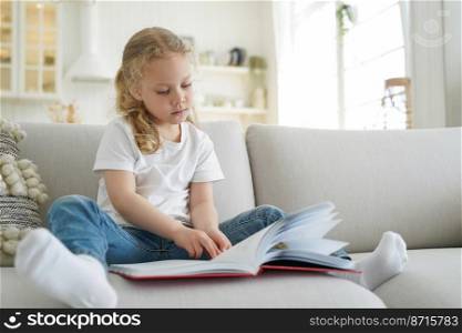 Pensive little child girl preschooler reading book, fairytale, sitting on couch in living room, small elementary kid daughter reads literature bestseller for children at home. Children’s education.. Little elementary child girl reads book, literature bestseller for children, sitting on sofa at home