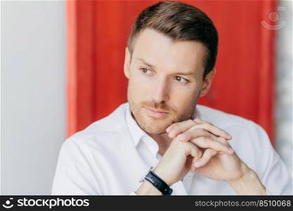 Pensive male director keeps hands presssed together, looks thoughtfully aside, being deep in thoughts before important meeting, poses indoor against red and white wall with copy space for your text