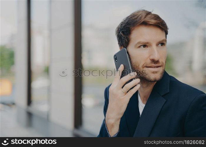 Pensive male entrepreneur focused into distance makes answering call uses mobile phone for communication holds mobile interview wears black formal jacket speaks to customer or business partner. Pensive male entrepreneur focused into distance makes answering call uses mobile phone