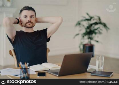 Pensive male freelancer takes break keeps hands behind head poses at desktop works on laptop computer has thoughtful expression thinks about future plans plans week dressed in black t shirt.