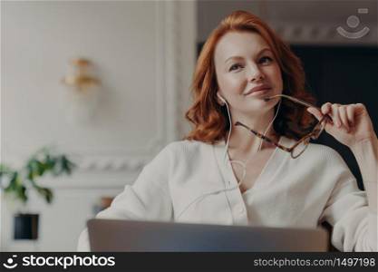 Pensive redhead adult woman listens music, takes break during freelance work, sits with laptop computer, has dreamy expression as enjoys lyrics song, holds spectacles, poses over home interior