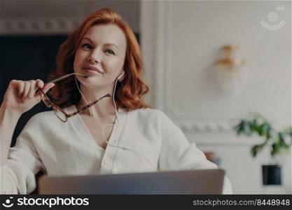 Pensive redhead adult woman listens music, takes break during freelance work, sits with laptop computer, has dreamy expression as enjoys lyrics song, holds spectacles, poses over home interior