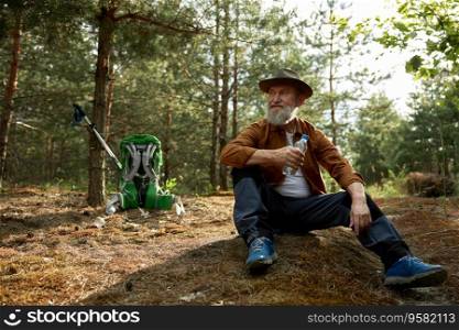 Pensive senior tourist drinking water from plastic bottle sitting on dried forest grass. Survival travel and hiking in woodland concept. Pensive senior tourist drinking water from plastic bottle while rest
