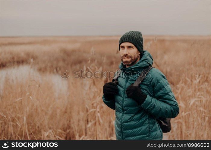 Pensive unshaven guy wears hat, green jacket and gloves, carries rucksack, poses against yellow autumn field background, has stroll outside, breathes fresh air, focused into distance. Tourism concept