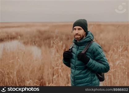 Pensive unshaven guy wears hat, green jacket and gloves, carries rucksack, poses against yellow autumn field background, has stroll outside, breathes fresh air, focused into distance. Tourism concept