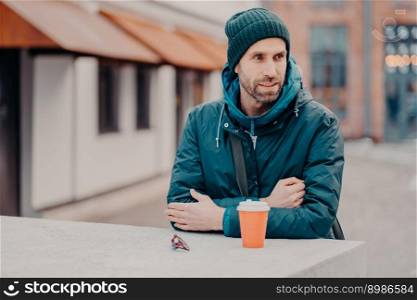 Pensive unshaven young Caucasian man with bristle, wears hat and jacket with hoody, drinks takeaway coffee, stands outdoor, has stroll. People, lifestyle, spare time and street style concept.