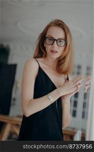 Pensive woman wearing stylish glasses and black dress looking at camera through mirror. Successful pretty female fashion model in eyewear posing for photo portrait. Eyeglasses online store advertising. Pensive female wearing stylish glasses, black dress, looking at camera. Eyeglasses store advertising