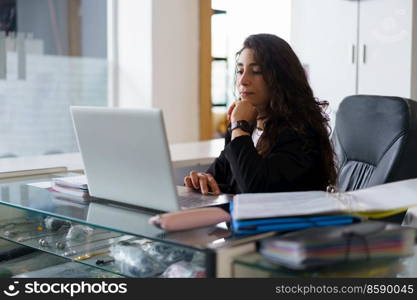 Pensive woman with wavy dark hair touching chin and browsing data on laptop while sitting at desk with sports supplies during work in gym. Thoughtful sports psychologist using netbook during work