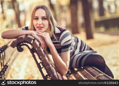 Pensive young blonde woman sitting on a bench in the street of a park with autumn colors. Beautiful girl in urban background wearing striped dress and black tights. Female with straight hair and blue eyes.