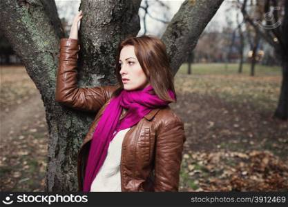 Pensive young brunette girl standing near tree in autumn park