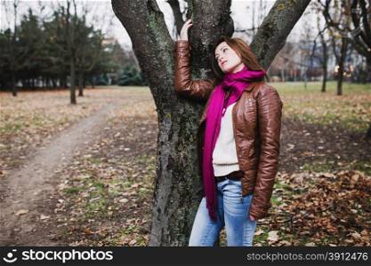 Pensive young brunette girl standing near tree in autumn park