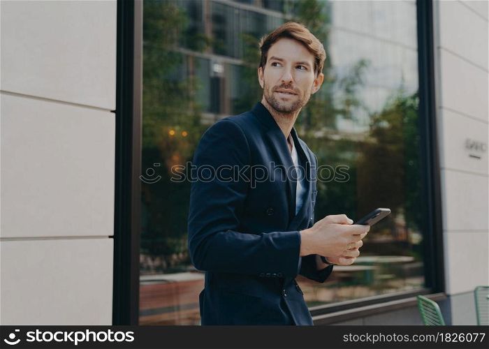 Pensive young businessman in dark suit holding smartphone, thinking about how to reply to partner, looking away while standing outside next to office building, selective focus on man with mobile phone. Businessman in dark suit holding smartphone and looking aside while walking city street