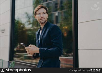 Pensive young businessman in dark suit holding smartphone, thinking about how to reply to partner, looking away while standing outside next to office building, selective focus on man with mobile phone. Businessman in dark suit holding smartphone and looking aside while walking city street