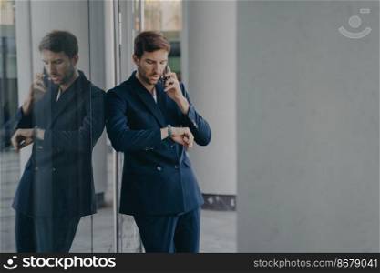 Pensive young businessman in formal suit talking on mobile phone standing outside of office center entrance while hastily glancing at his hand watch, worried about being on time for company meeting. Pensive businessman in suit talking on mobile phone standing outside, looking at his hand watch