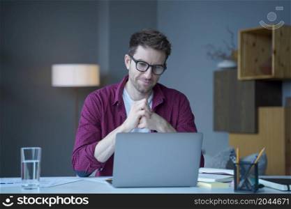 Pensive young German man home office employee sits at desk at workplace operates online on laptop pondering thinking. Focused positive male looks at computer screen browsing internet on modern gadget. Pensive young German man home office employee sits at desk at workplace operates online on laptop