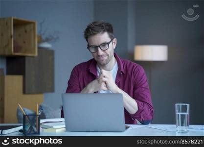 Pensive young German man home office employee sits at desk at workplace operates online on laptop pondering thinking. Focused positive male looks at computer screen browsing internet on modern gadget. Pensive young German man home office employee sits at desk at workplace operates online on laptop