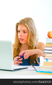 Pensive young girl sitting at table with books and looking on laptop&#xA;