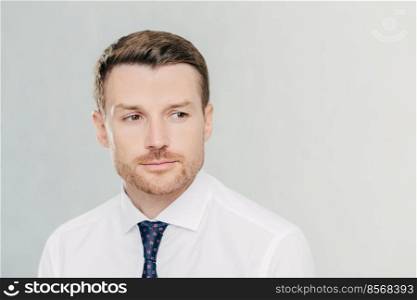 Pensive young male businessman looks thoughtfully aside, thinks about business project, focused into distance, isolated over white background with copy space aside for advertisement or text.