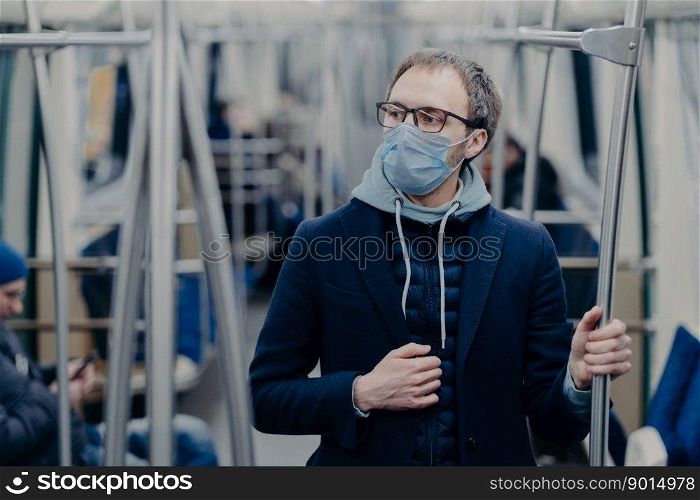Pensive young man in eyewear wears protective surgical mask during coronavirus outbreak, poses in public transportation, thinks how to overcome disease. Virus protection, quarantine concept.