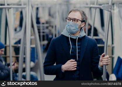 Pensive young man in eyewear wears protective surgical mask during coronavirus outbreak, poses in public transportation, thinks how to overcome disease. Virus protection, quarantine concept.