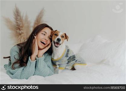 Peop≤and animals concept. Joyful pretty young woman express love to dog, spends≤isure time with pet, lie to≥ther on bed, looks tenderly at animal, feels not bored with true loyal friend