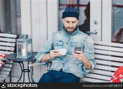 Peop≤,≤isure and technology concept. Positive bearded young ma≤recreats at terrace cafe, holds modern smart pho≠, messa≥s onli≠with friends, drinks tea or coffee, wears denim jacket