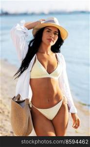 peop≤,∑mer holidays and≤isure concept - happy woman in bikini swimsuit, white shirt and straw hat with bag walking along beach. happy woman in bikini and shirt walking on beach