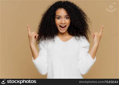People, advertisement and emotions concept. Glad young female points upwards with index fingers, shows awesome item on sale, wears white sweater, exclaims happily, isolated on brown studio wall