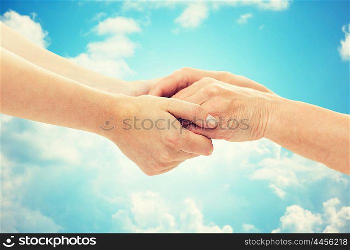 people, age, family, care and support concept - close up of senior woman and young woman holding hands over blue sky and clouds background