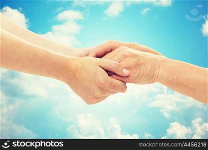 people, age, family, care and support concept - close up of senior woman and young woman holding hands over blue sky and clouds background
