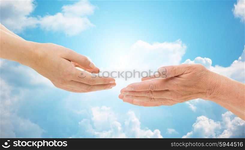 people, age, family, care and support concept - close up of senior woman and young woman reaching hands out to each other over blue sky and clouds background