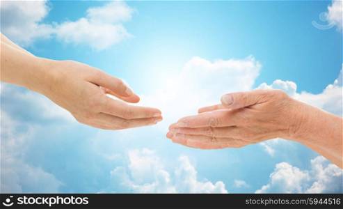 people, age, family, care and support concept - close up of senior woman and young woman reaching hands out to each other over blue sky and clouds background
