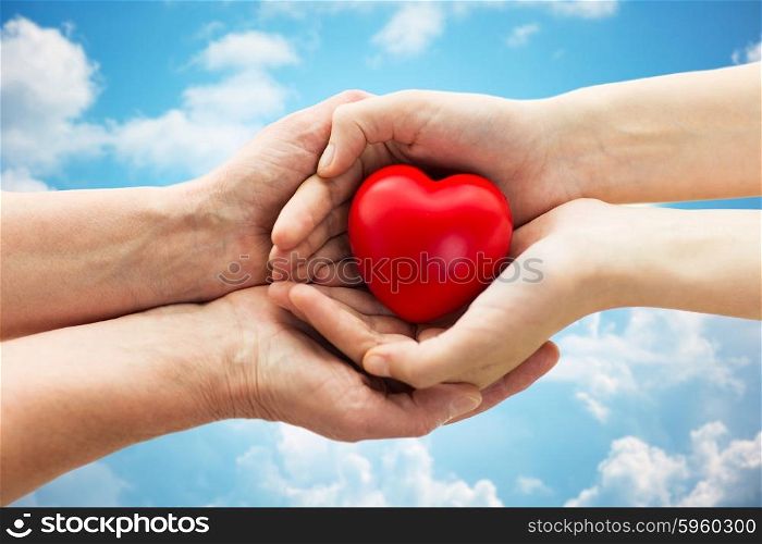 people, age, family, love and health care concept - close up of senior woman and young woman hands holding red heart over blue sky and clouds background