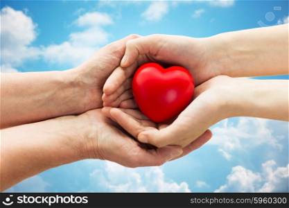 people, age, family, love and health care concept - close up of senior woman and young woman hands holding red heart over blue sky and clouds background