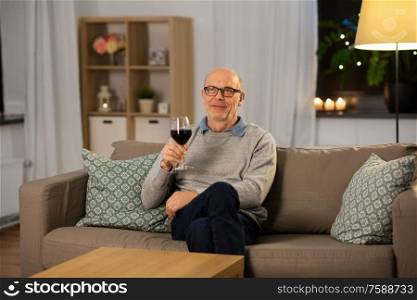 people, alcohol and drinks concept - happy senior man drinking red wine from glass at home in evening. senior man drinking red wine from glass at home