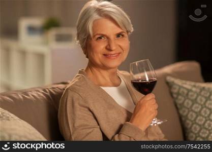 people, alcohol and drinks concept - happy smiling senior woman drinking red wine from glass at home in evening. senior woman drinking red wine at home in evening