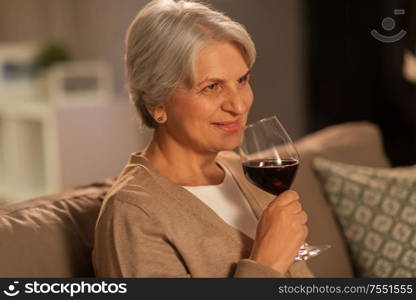 people, alcohol and drinks concept - happy smiling senior woman drinking red wine from glass at home in evening. senior woman drinking red wine at home in evening