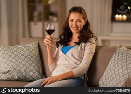 people, alcohol and drinks concept - happy young woman drinking red wine from glass at home in evening. young woman drinking red wine at home in evening