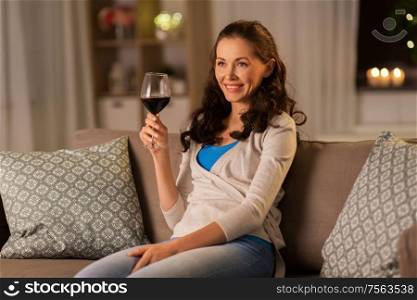 people, alcohol and drinks concept - happy young woman drinking red wine from glass at home in evening. young woman drinking red wine at home in evening