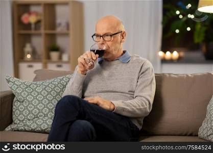 people, alcohol and drinks concept - senior man drinking red wine from glass at home in evening. senior man drinking red wine from glass at home