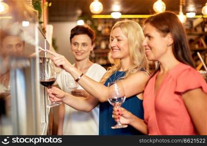 people, alcohol and lifestyle concept - happy women pouring red wine from dispenser at bar or restaurant. happy women pouring wine from dispenser at bar