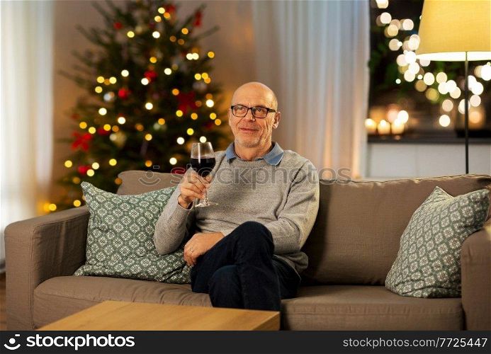 people, alcohol and winter holidays concept - happy senior man drinking red wine from glass at home in evening over christmas tree lights on background. old man drinking red wine from glass on christmas